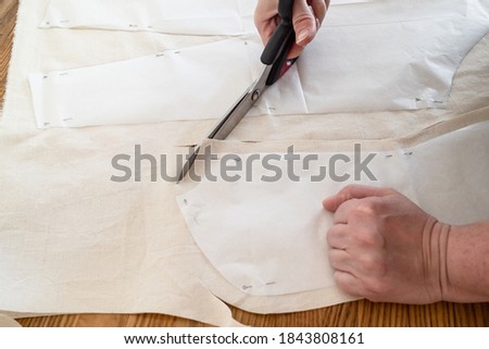 designer's hand cuts calico cloth by scissors according with paper layouts of dress on wooden table at home Royalty-Free Stock Photo #1843808161
