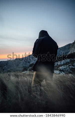 A man with a black jacket stands in a field of reed at the mountains while waiting the perfect moment to take a picture of a landscape during sunrise