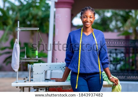 Beautiful African seamstress standing in front of sewing machine with measuring tape around neck,cheerfully looking at camera-concept on millennial black female entrepreneurship  Royalty-Free Stock Photo #1843937482