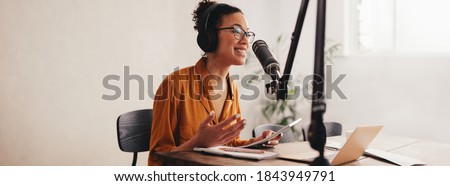 Woman recording a podcast on her laptop computer with headphones and a microscope. Female podcaster making audio podcast from her home studio. Royalty-Free Stock Photo #1843949791