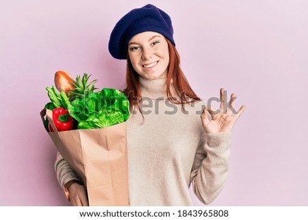 Young red head girl holding paper bag with bread and groceries doing ok sign with fingers, smiling friendly gesturing excellent symbol 