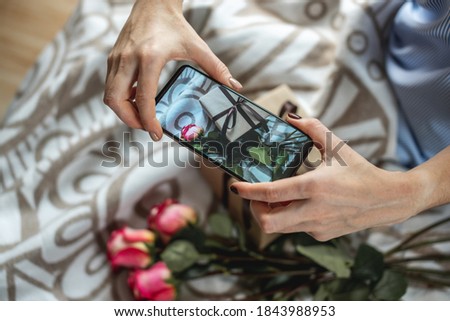 A woman's hands are holding a mobile phone and taking photos of flowers and a gift box on a bedspread. Concept of taking photos on the phone and a birthday or other holiday.