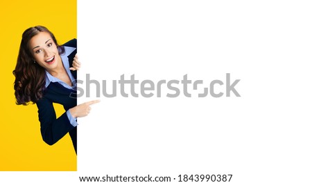 Happy excited woman in black confident suit and light blue blouse showing pointing blank banner signboard. Business and advertising concept. Copy space place for text. Yellow orange color background.