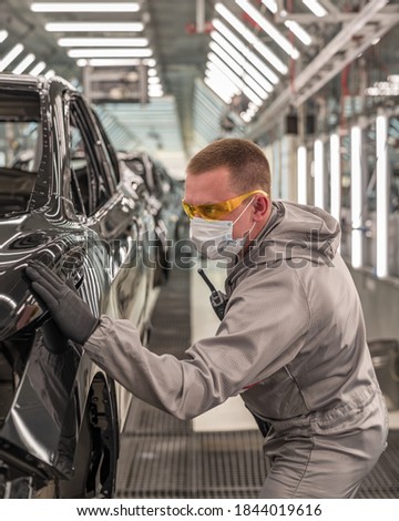 An employee of the paint shop of an automobile factory in a medical mask on his face checks the quality of the painted surface. Working during a pandemic