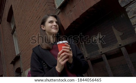 Portrait of young attractive woman standing along red brick wall of old  living house, drinking coffee outdoor, looking at the street, dutch angle Royalty-Free Stock Photo #1844056465