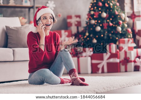 Photo of charming young girl sit carpet hold phone talk explain wear santa cap red sweater jeans socks decorated living room indoors
