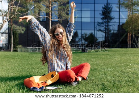 Happy young blonde hipster woman student with backpack sitting on grass at university.