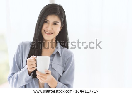 Young and beautiful Asian woman holding white ceramic coffee cup with relax and easy gesture while looking to camera. There is soft curtain in background. Idea for happy in the morning time.