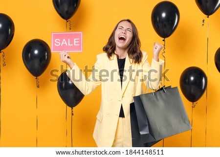 Happy young woman wearing suit jacket hold package bag with purchases after shopping sign with SALE title doing winner gesture on yellow background with air balloons studio portrait. Black friday