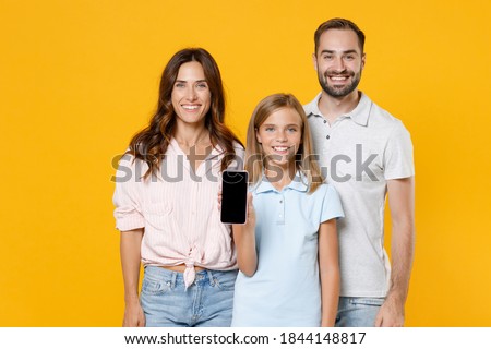 Smiling young parents mom dad with child kid daughter teen girl in basic t-shirts hold mobile phone with blank empty screen mock up copy space isolated on yellow background studio. Family day concept