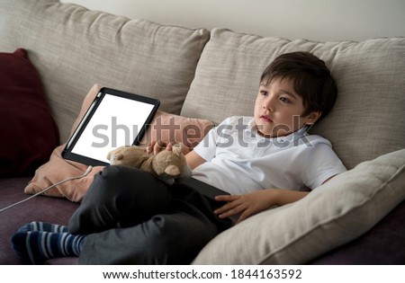 Happy boy watching  cartoon while waiting for Tablet charging,  School Kid sitting alone on sofa holding remote control and watching TV, Child relaxing in living room after back from school 