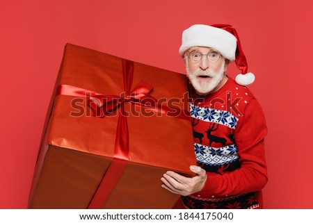 Excited senior man in santa hat looking at camera while holding present isolated on red