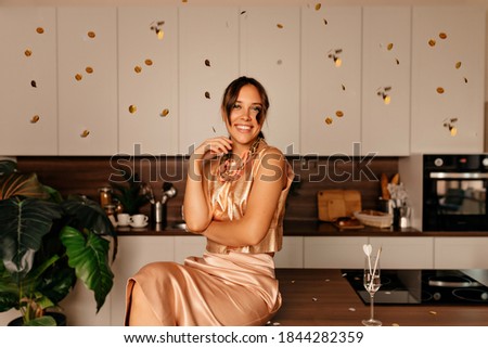 Happy exited woman in shine bright dress sitting ay the kitchen and celebrating birthday, New Year with confetti