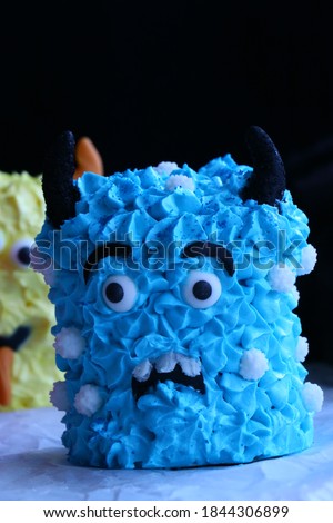 Close-up picture of a cute blue mini monster cake, part of yellow monster cake blurry visible in the background. Decorated for Halloween.