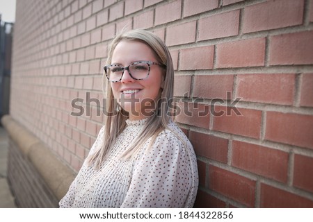 Beautiful, Young 30 Years Old Girl with Blonde Hair, in Glasses, wearing white dress, Smiling and Posing for camera, red Bricks background, Happy Woman  