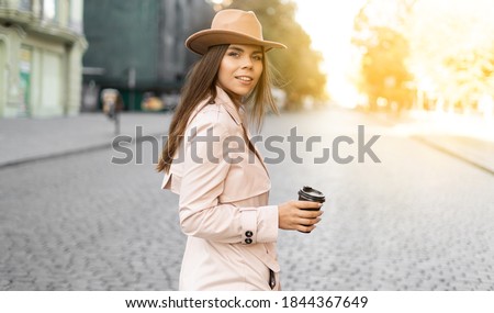 young woman walks through the city streets dressed in a hat and a raincoat. Coffee to go.
