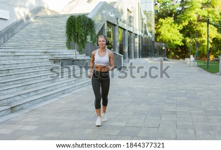 Young attractive Caucasian blonde woman in sportswear doing morning jogging while listening to music in headphones outdoors in the city. Morning workout, healthy lifestyle