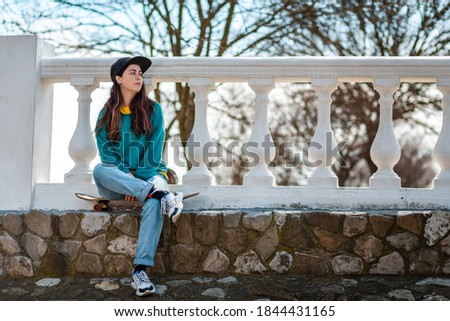 A beautiful hipster teenage girl sitting relaxed on a skateboard and looking into the distance. In the background is a White balustrade. Copy space. Concept of sports lifestyle and street culture