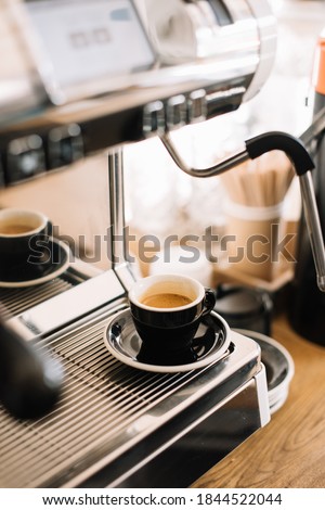 Delicious fresh morning espresso coffee in a black ceramic cup with a saucer, standing on a metal tray of the coffee machine, vertical photo
