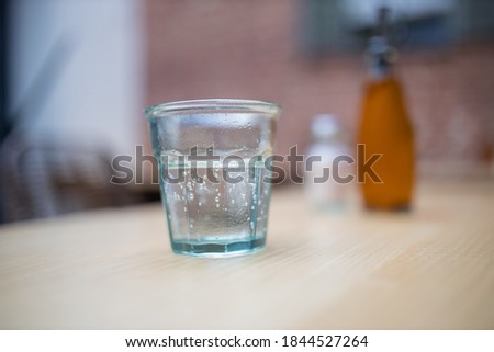 Close up picture of a glass of water on a wooden table with a blurry brown bottle and the interior of a restaurant as a background
