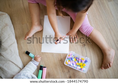 Landscape picture of a little girl in pink clothing sitting on a wooden floor and doing sums on a paper sheet with a pink textmarker and colorful cotton balls alongside her baby sibling