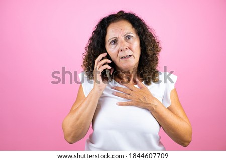 Middle age woman wearing casual white shirt standing over isolated pink background talking on the phone with a worried expression