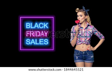 Surprised pinup woman eating heart shape lollipop. Girl in pin up cloth. Blond model at retro fashion and vintage concept. Dark background. Black Friday sales neon light sign. 