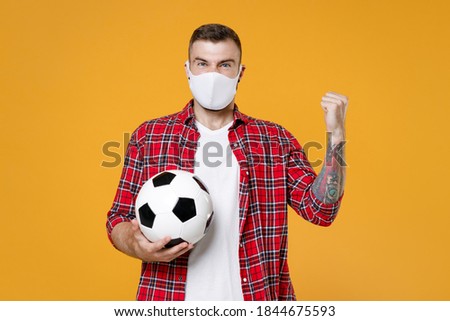 Angry man football fan in face mask to safe from coronavirus virus covid-19 cheer up support favorite team with soccer ball clenching fists isolated on yellow background. People sport leisure concept