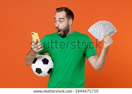 Shocked young man football fan in green t-shirt cheer up support favorite team with soccer ball using mobile cell phone fan of cash money isolated on orange background. People sport leisure concept
