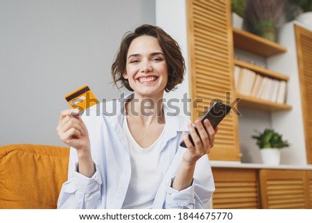 Smiling young woman in casual clothes sit on couch spending time in living room at home. Rest relax good mood leisure lifestyle concept. Mock up copy space. Using mobile phone, hold credit bank card
