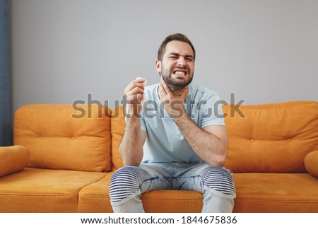 Sick exhausted young bearded man in casual blue t-shirt hold paper napkin coughing sneezing put hand on neck throat keeping eyes closed sitting on couch resting spending time in living room at home