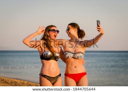 Mother and daughter make selfie on beach smiling and making funny faces. Mother and daugher teen brunettes are taking pictures with mobile phones on the beach.
