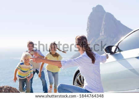 Kids running towards their mother as father clicks their picture on a hill near the sea beside a parked car