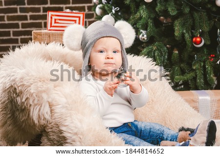 Subject children christmas new year. Caucasian little funny baby boy 1 year old sitting sleigh bear skin Christmas tree head warm hat with balabons balls, balls and jeans around box gifts in evening.