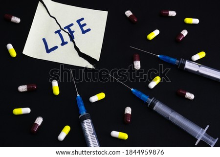 Danger of drugs to life concept. Torn sheet of paper with inscription "Life" on black background with used syringes and many pills