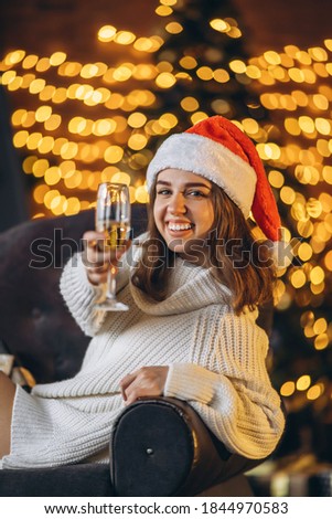 Christmas, New Year. Pretty woman in warm sweater, socks and Christmas hat, sitting on the chair with champagne and new year tree and lights behind