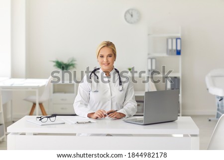 Friendly young woman, working as doctor, sitting at desk in hospital office, smiling and looking at camera. Portrait of medical worker for health vlog, video call picture, consultation with patient