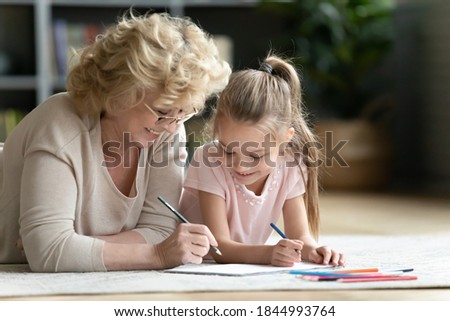Head shot caring loving older granny or nanny lying on floor with cute small kid girl, drawing pictures in paper album. Affectionate elderly woman teaching painting little preschool granddaughter.