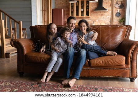 Happy young Caucasian family with two small kids sit relax on sofa at home watch movie on smartphone. Smiling parents with little children look at cell screen talk speak on video call on cellphone.