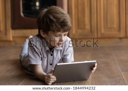Cute little 7s Caucasian boy lying on warm floor at home look at tablet screen watch video or cartoon online. Happy smart small kid child have fun using modern pad device, play video game on gadget.