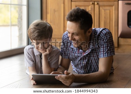 Smiling young Caucasian dad and small son have fun play online game on modern tablet gadget at home. Happy father teach little boy child use pad device, watch video or cartoon. Technology concept.
