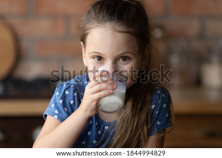 Portrait of little Caucasian girl drink tasty nutritional wholegrain milk from glass at home. Smiling small kid child enjoy delicious organic healthy lactose free yoghurt. Diet, healthcare concept.