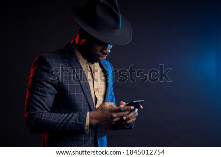 Portrait of confident stylish young african man in formalwear, holding mobile phone, looking at screen, standing against black background. Successful businessman. Business, technology.