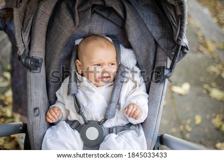 Picture of a distressed baby in a gray stroller looking at the distance and crying with the sidewalk covered in autumn leaves as background