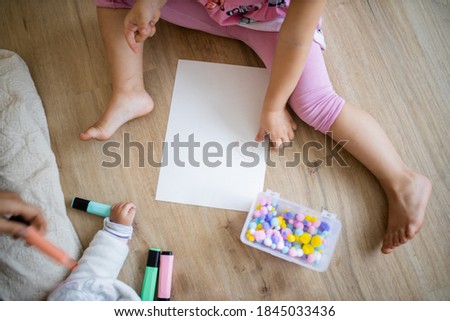 Landscape picture of a little girl in pink clothing sitting on a wooden floor with a white paper sheet, alongside green and pink textmarkers, colorful cotton balls, and her baby sibling