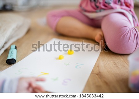 Picture of the legs of a little girl sitting on a wooden floor alongside two yellow cotton balls, sums on a paper sheet, and the blurry arm of a baby