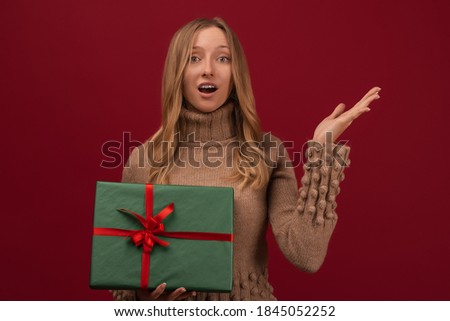 Image of charming young blonde womanin trendy knitted sweater smiling and holding gift with red ribbon. Studio shot red background. New Year Women's Day birthday holiday concept