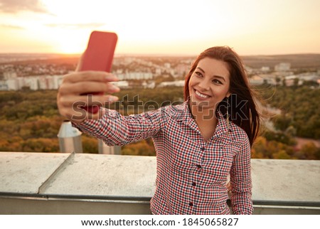 Joyful young female in casual clothes taking selfie on mobile phone while standing on rooftop terrace of urban building at sunset time in summer evening