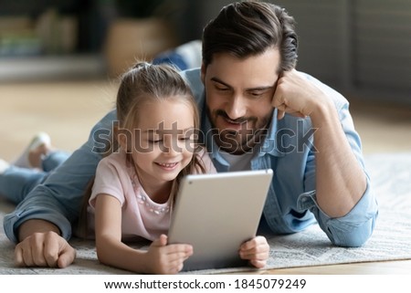 Caring young father cuddling small 6 years old daughter, lying together on floor using digital tablet at home. Happy little cute kid girl relaxing with dad, watching funny cartoons on gadget.