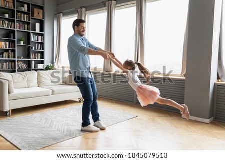 Happy small 7 years old funny girl holding daddy's hands, flying in air in living room. Carefree smiling young father twisting little preschool kid daughter, entertaining having fun together at home. Royalty-Free Stock Photo #1845079513